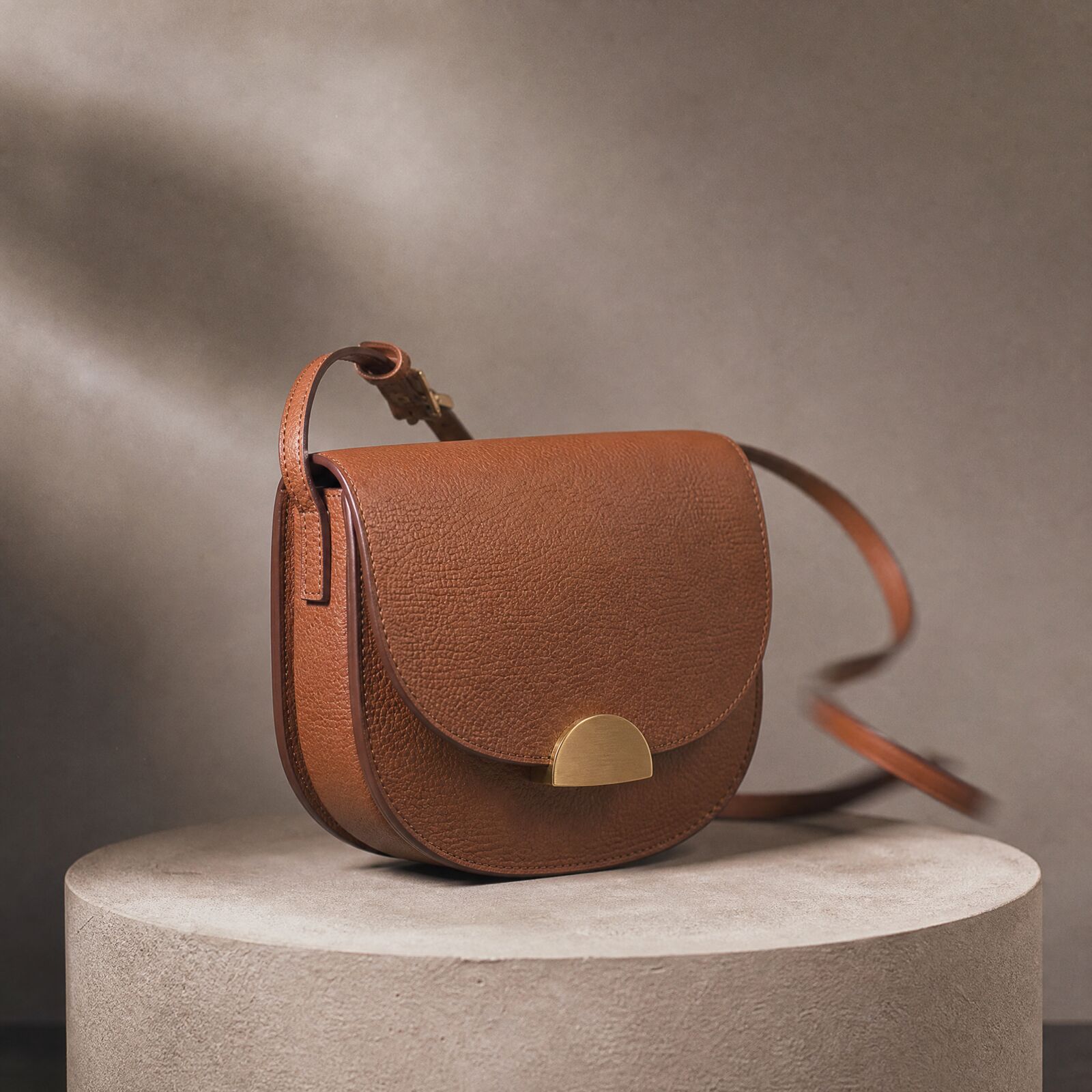 A rounded tan leather saddle bag on a stone plinth in warm raking light against a beige wall.